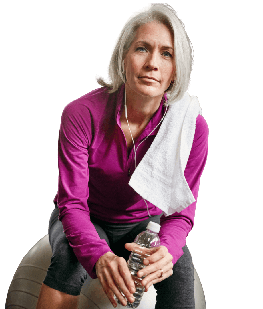 Eine Person mit grauem Haar sitzt auf einem Fitnessball, trägt ein lila Oberteil, hält eine Wasserflasche und hat ein Handtuch über der Schulter. Sie trägt Ohrhörer und blickt geradeaus.